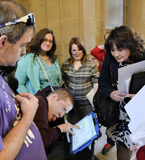 A young man using a wheelchair and communication device talks with a female legislator as his peers watch.