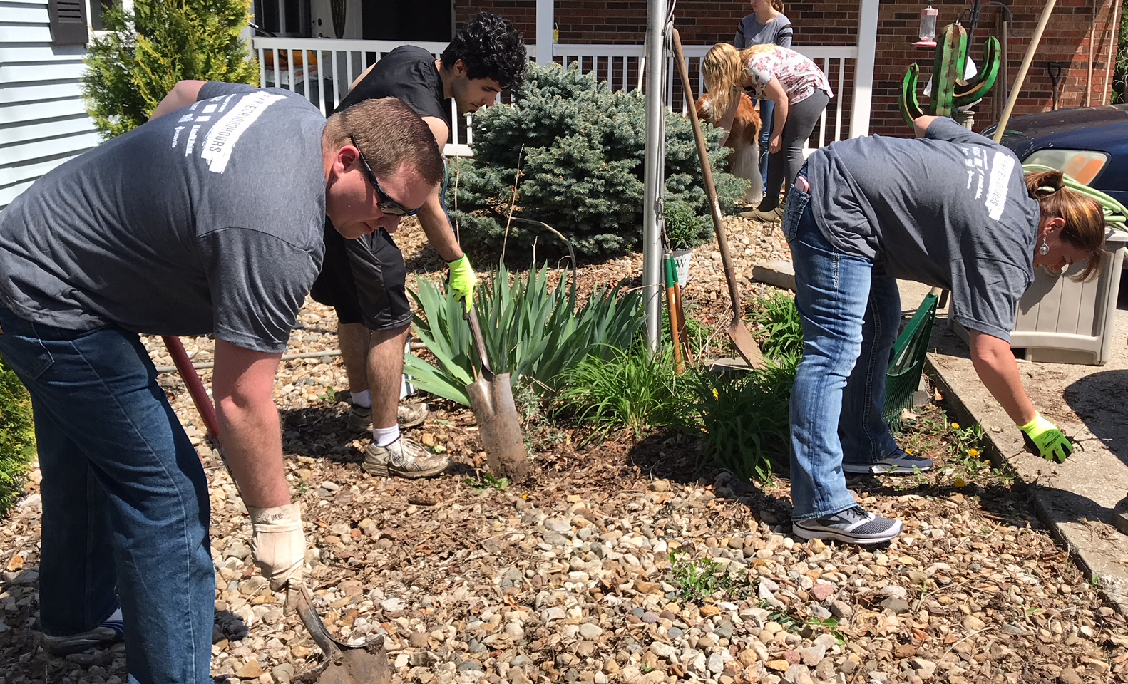 Volunteers in matching shirts use shovels and hand tools to clean up mulch and landscaping at a group home.