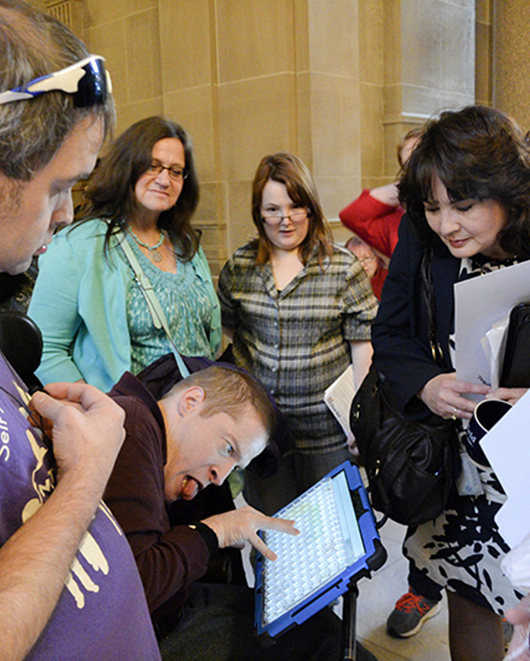 A young man using a wheelchair and communication device talks with a female legislator as his peers watch.